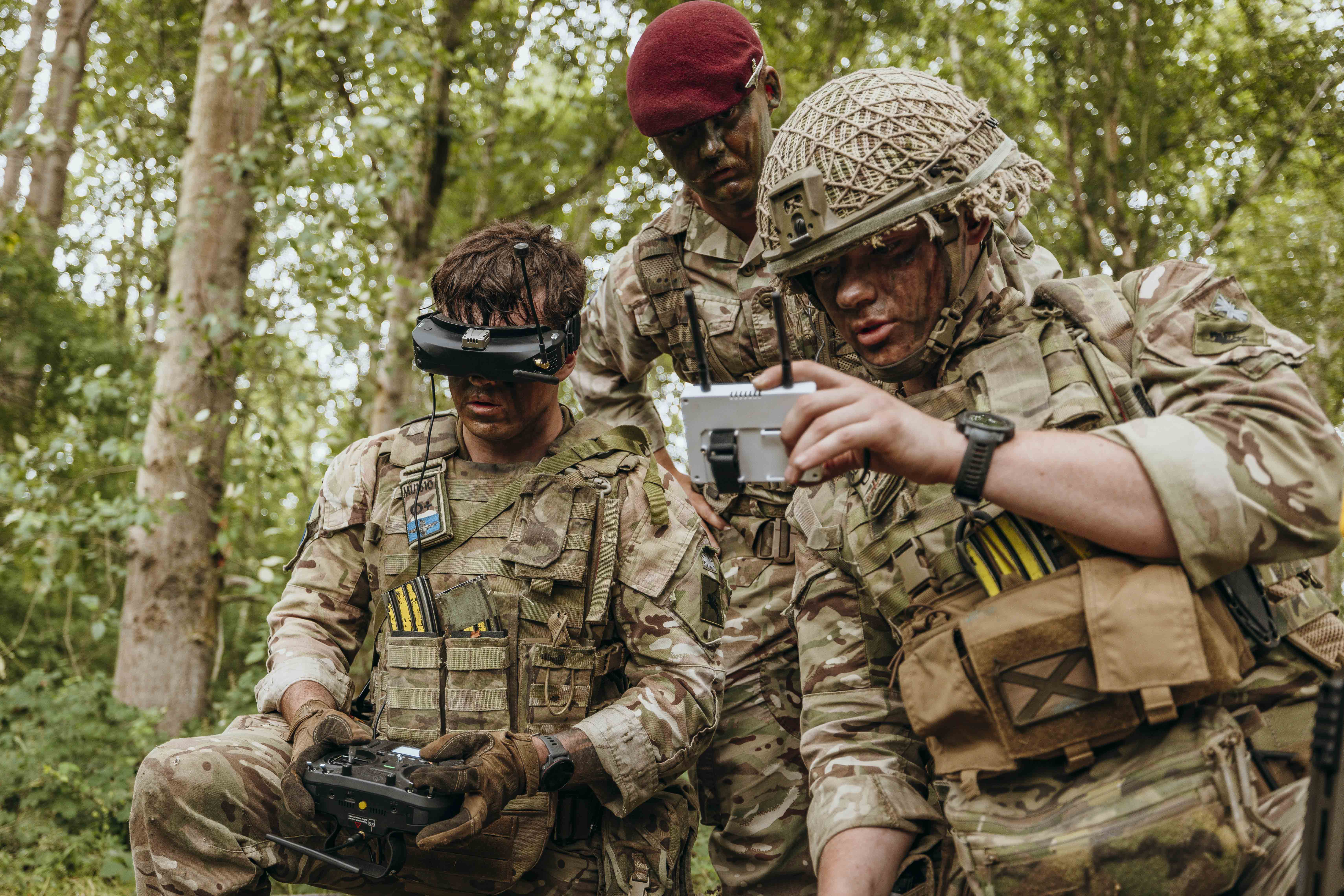 Three soldiers in camouflage gear operate a drone controller and monitor its feed in a dense forest setting.