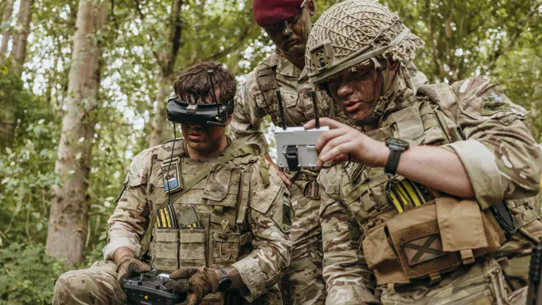 Three soldiers in camouflage gear operate a drone controller and monitor its feed in a dense forest setting.