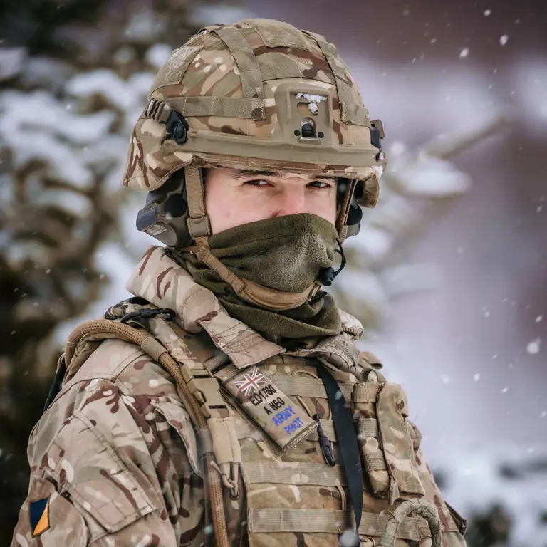 A portrait of a soldier holding a camera. He is wearing body armour and helmet. It is snowing.