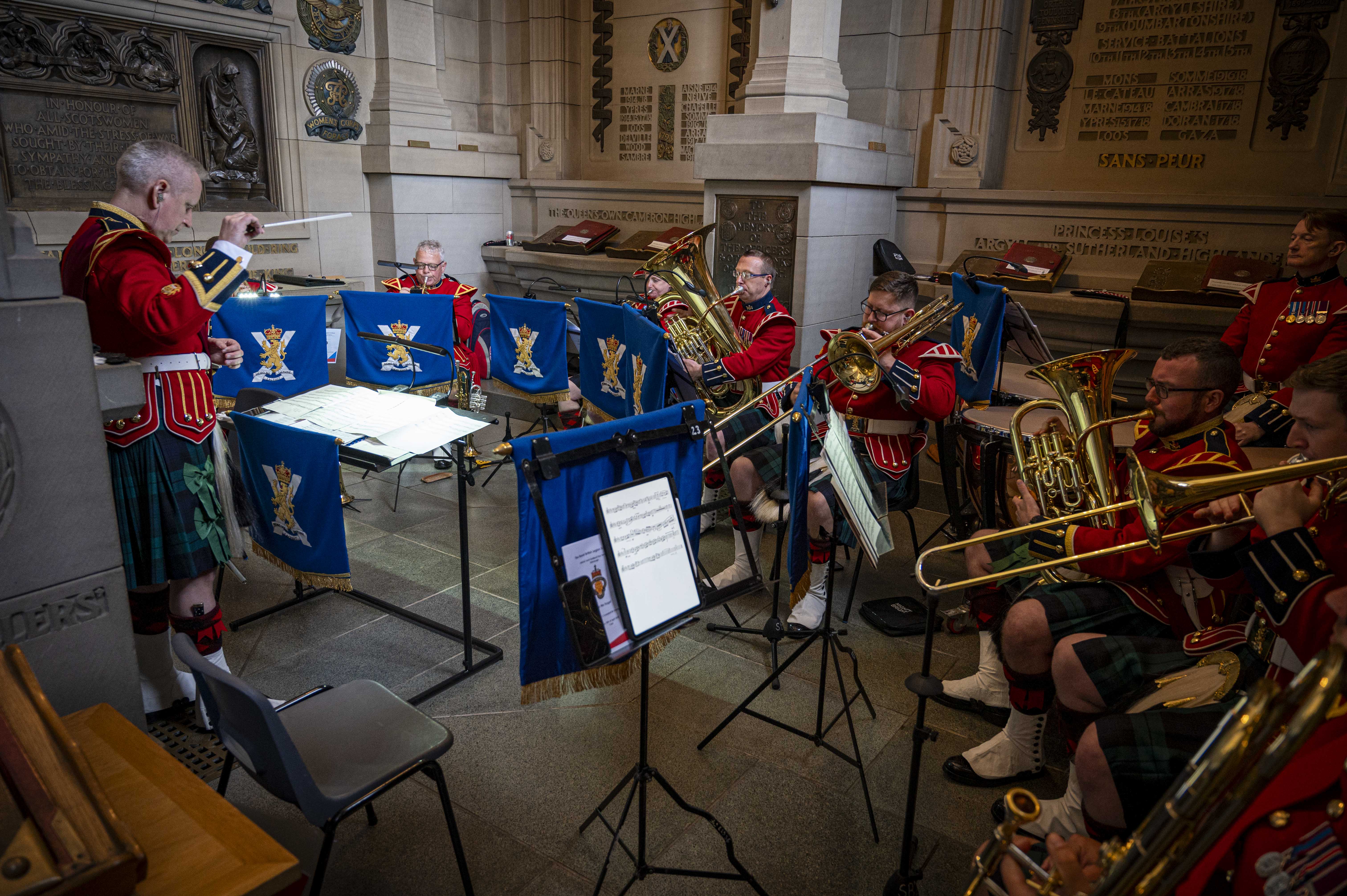 A musical ensemble in red jackets and kilts performs indoors, surrounded by ornate architectural details. Each member holds a brass instrument, and music stands are in front of them. There are music sheets on some stands. The setting appears to be historical, with rich decorations on the walls. The conductor stands to the left, guiding the musicians.
