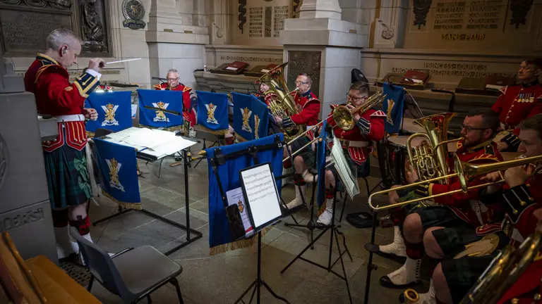 A musical ensemble in red jackets and kilts performs indoors, surrounded by ornate architectural details. Each member holds a brass instrument, and music stands are in front of them. There are music sheets on some stands. The setting appears to be historical, with rich decorations on the walls. The conductor stands to the left, guiding the musicians.