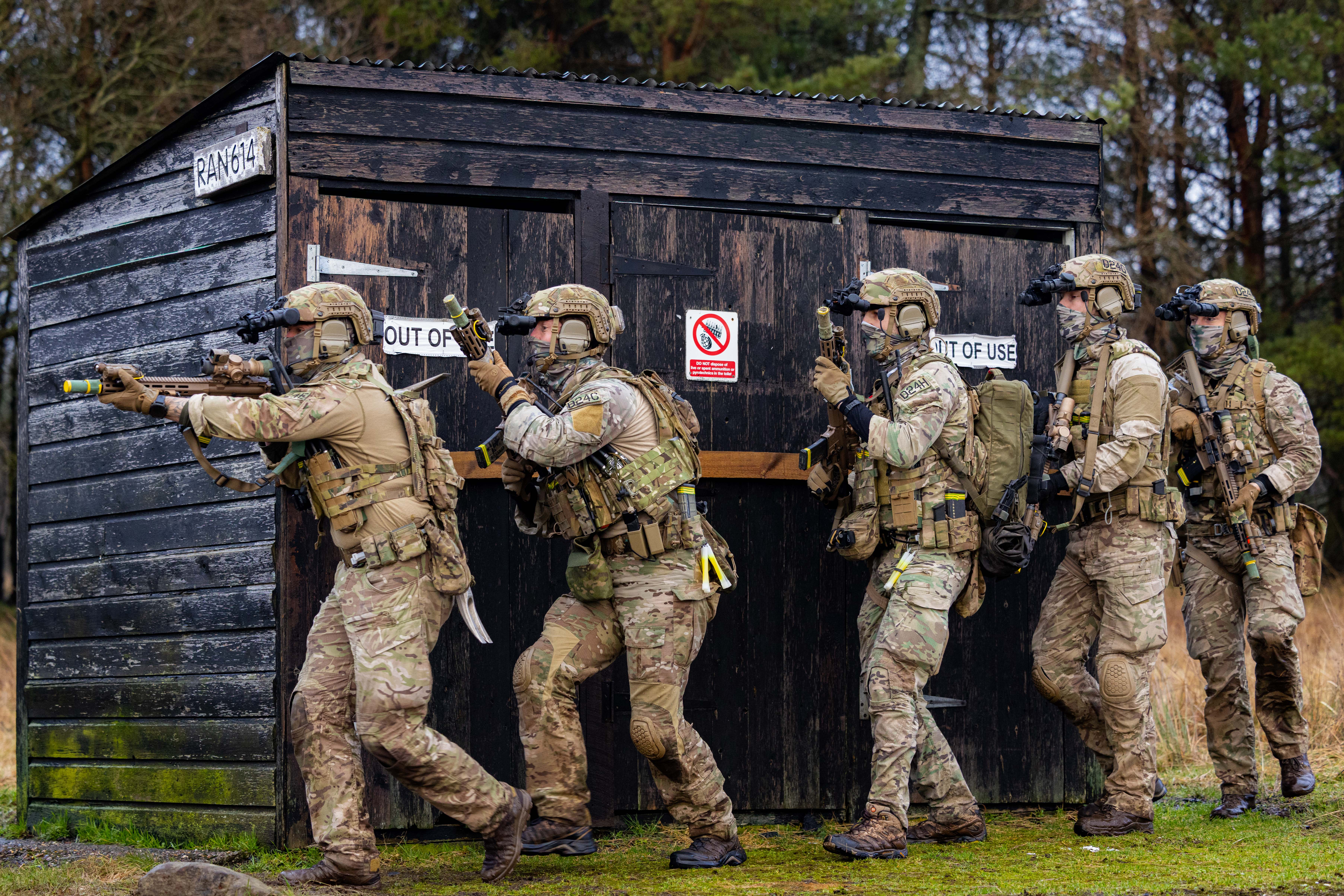 Five soldiers in tactical gear advancing cautiously beside a black wooden structure labeled and 'OUT OF USE'.