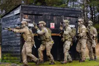 Five soldiers in tactical gear advancing cautiously beside a black wooden structure labeled and 'OUT OF USE'.