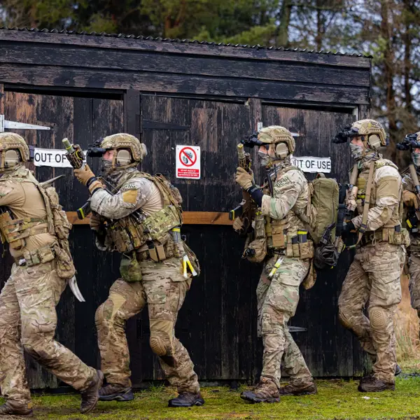 Five soldiers in tactical gear advancing cautiously beside a black wooden structure labeled and 'OUT OF USE'.