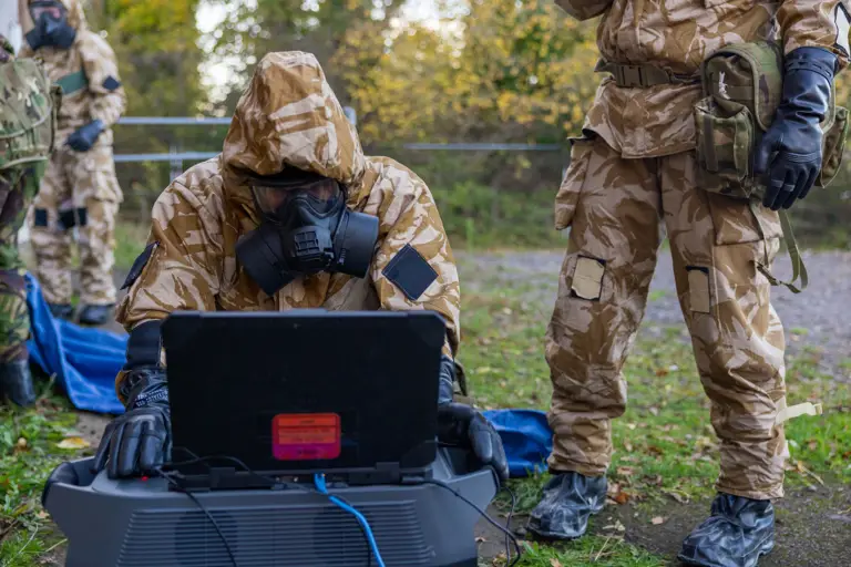 Soldier in camouflage and gas mask operating a laptop outdoors during a field exercise.