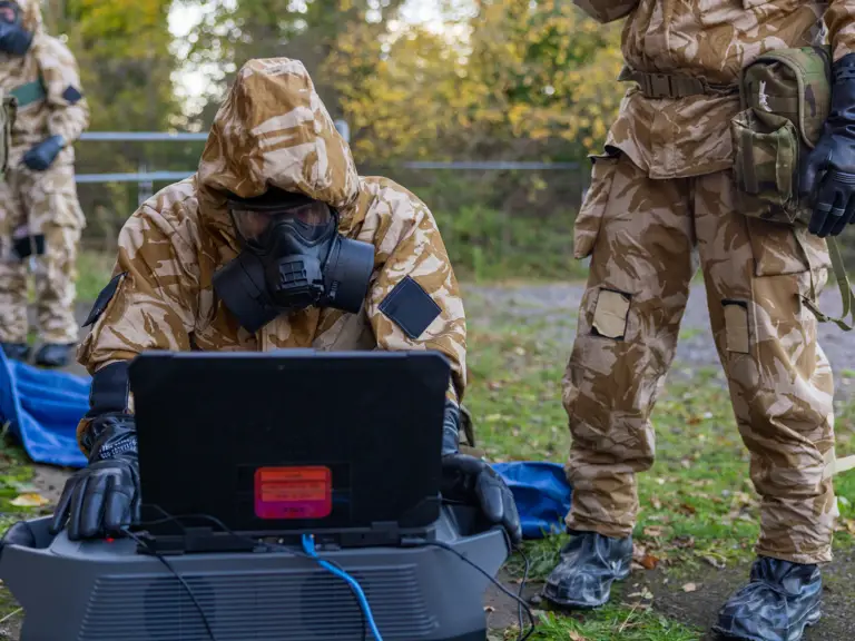 Soldier in camouflage and gas mask operating a laptop outdoors during a field exercise.