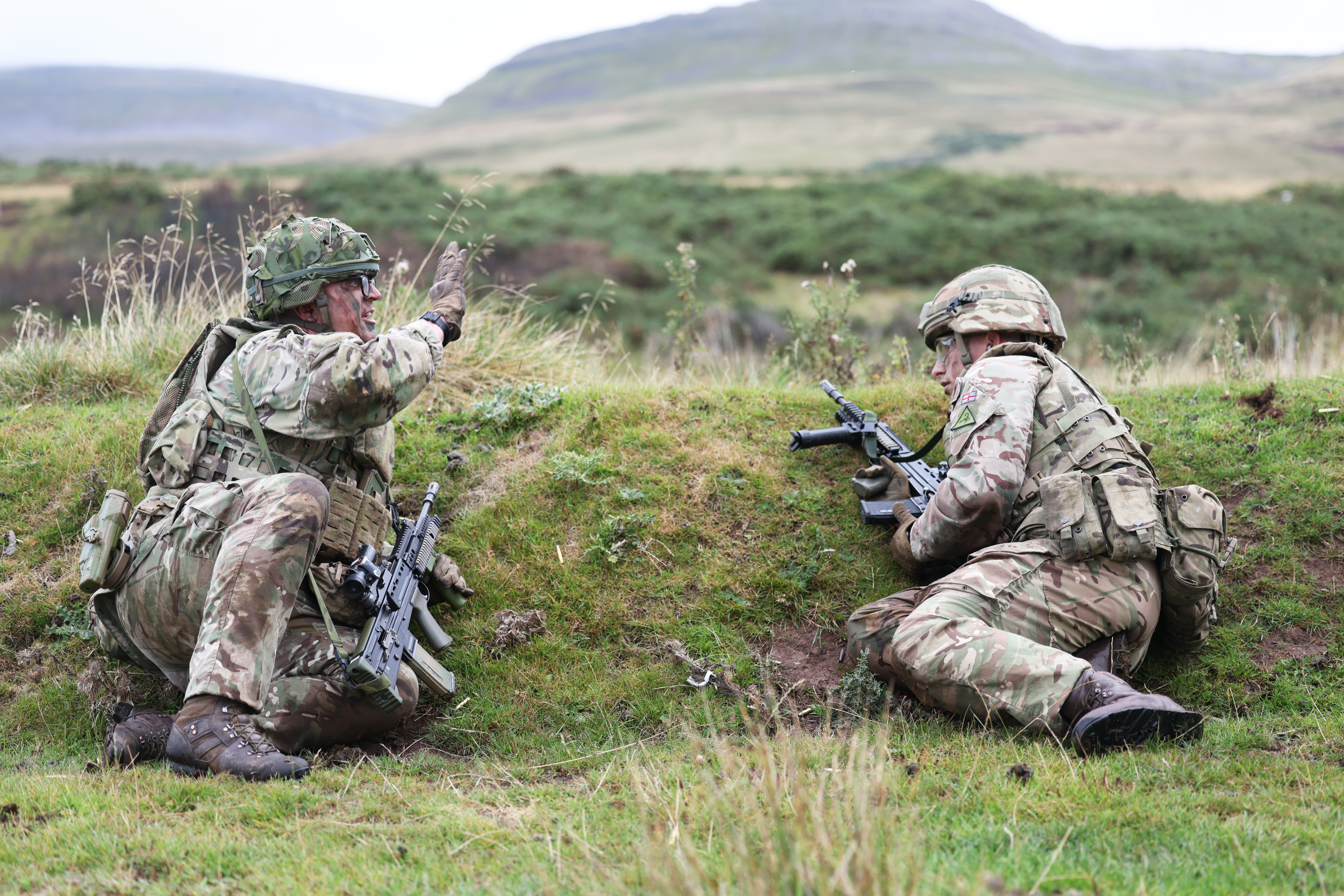 Two soldiers in camouflage gear crouch behind a grassy embankment during a training exercise in a hilly outdoor area.