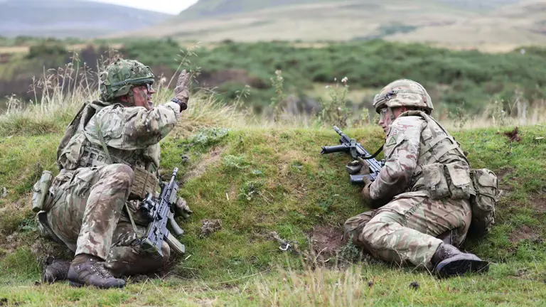 Two soldiers in camouflage gear crouch behind a grassy embankment during a training exercise in a hilly outdoor area.