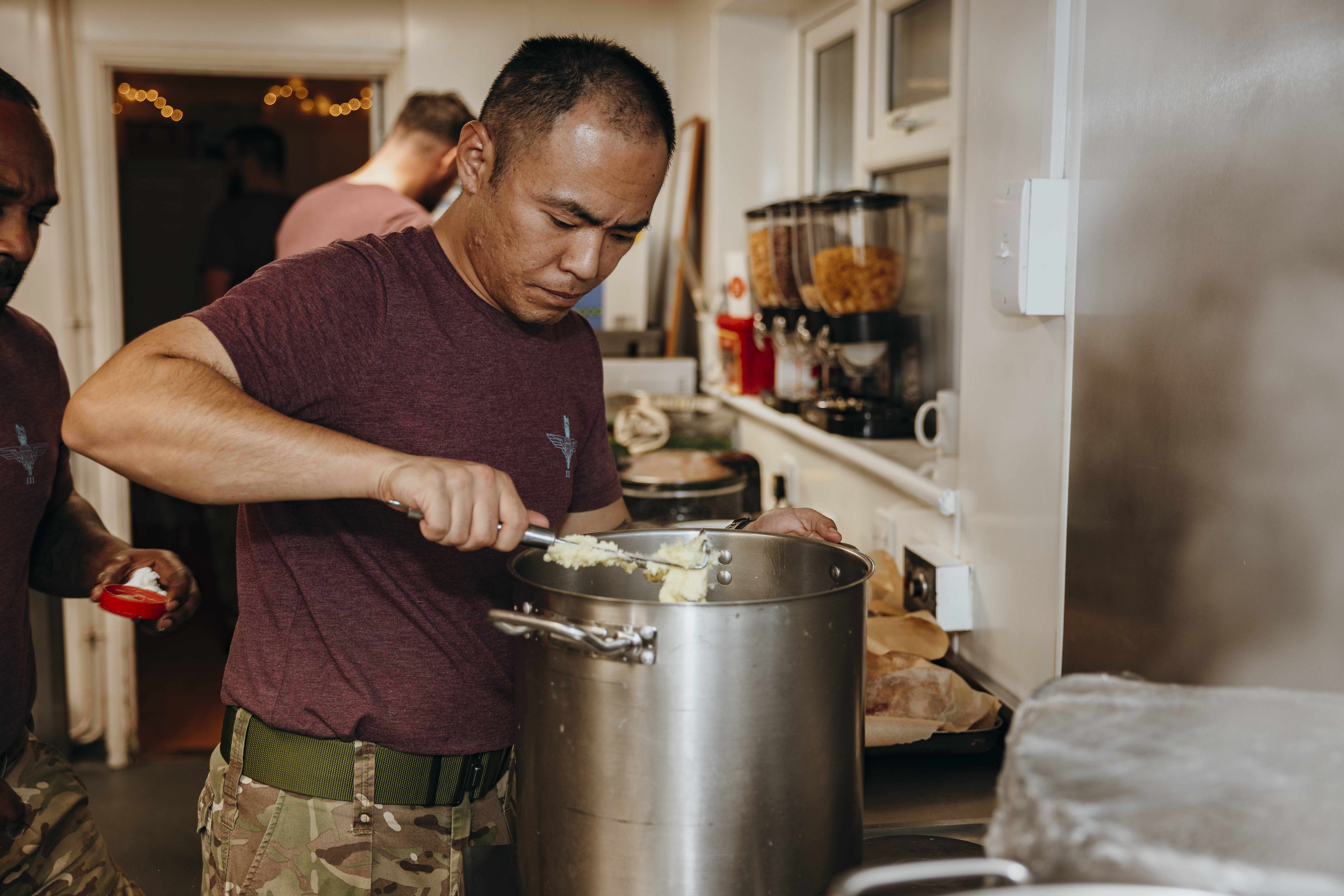 A man in a purple t-shirt and combat trousers prepares mashed potato. 