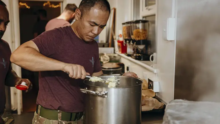 A man in a purple t-shirt and combat trousers prepares mashed potato.
