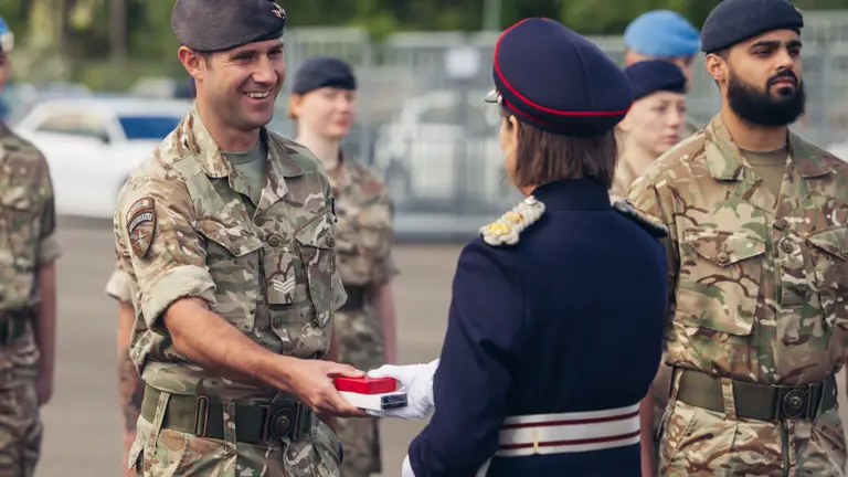 A soldier in uniform smiles as a woman in ceremonial attire hands him a medal.