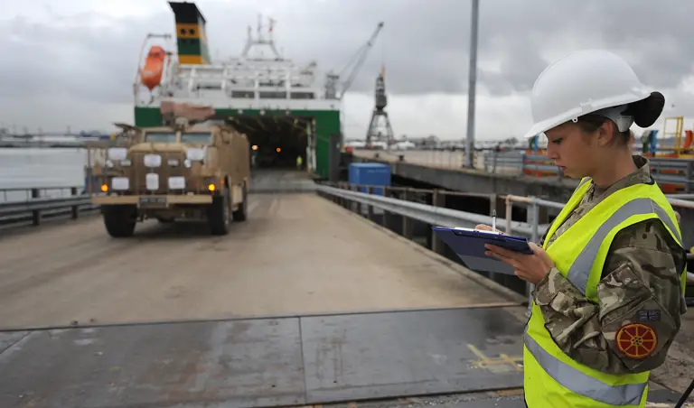 A movement controller makes notes on a clipboard as a military vehicles drives off a ship.