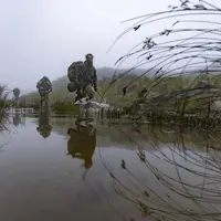 Military personnel wade through a shallow water body, surrounded by misty, rugged terrain. They carry gear and wear camouflage, indicating a tactical operation. Grasses and rocks flank the water, reflecting the soldiers' movements.
