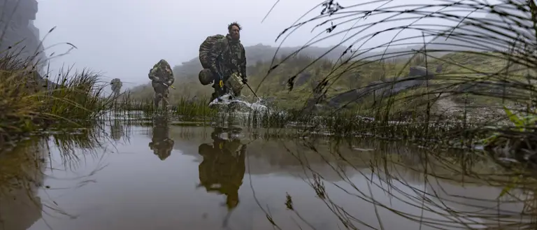 Military personnel wade through a shallow water body, surrounded by misty, rugged terrain. They carry gear and wear camouflage, indicating a tactical operation. Grasses and rocks flank the water, reflecting the soldiers' movements.