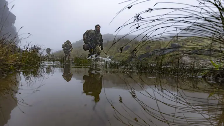 Military personnel wade through a shallow water body, surrounded by misty, rugged terrain. They carry gear and wear camouflage, indicating a tactical operation. Grasses and rocks flank the water, reflecting the soldiers' movements.