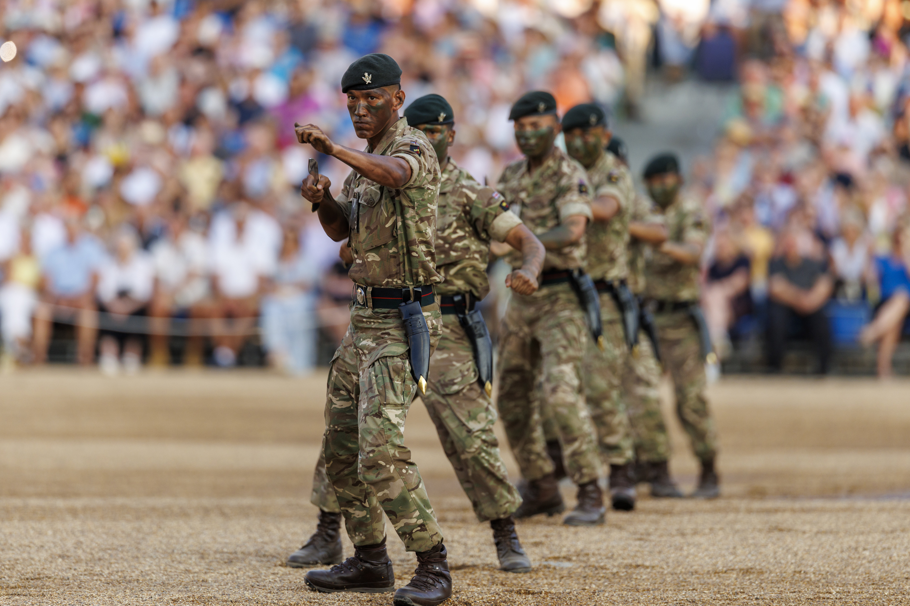 Military personnel in camouflage uniforms perform a coordinated display, holding knives, in front of a large audience. The scene is set in a stadium or similar venue.