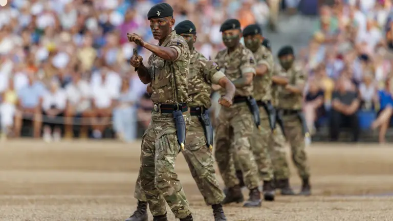 Military personnel in camouflage uniforms perform a coordinated display, holding knives, in front of a large audience. The scene is set in a stadium or similar venue.