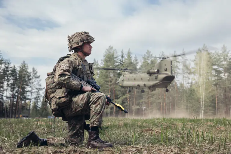 Soldier in camouflage uniform, helmet and holding a rifle is shown taking a knee as a Boeing Chinook takes flight over his head. Their is a woodland backdrop.