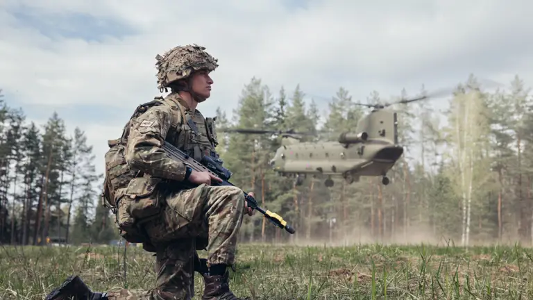 Soldier in camouflage uniform, helmet and holding a rifle is shown taking a knee as a Boeing Chinook takes flight over his head. Their is a woodland backdrop.