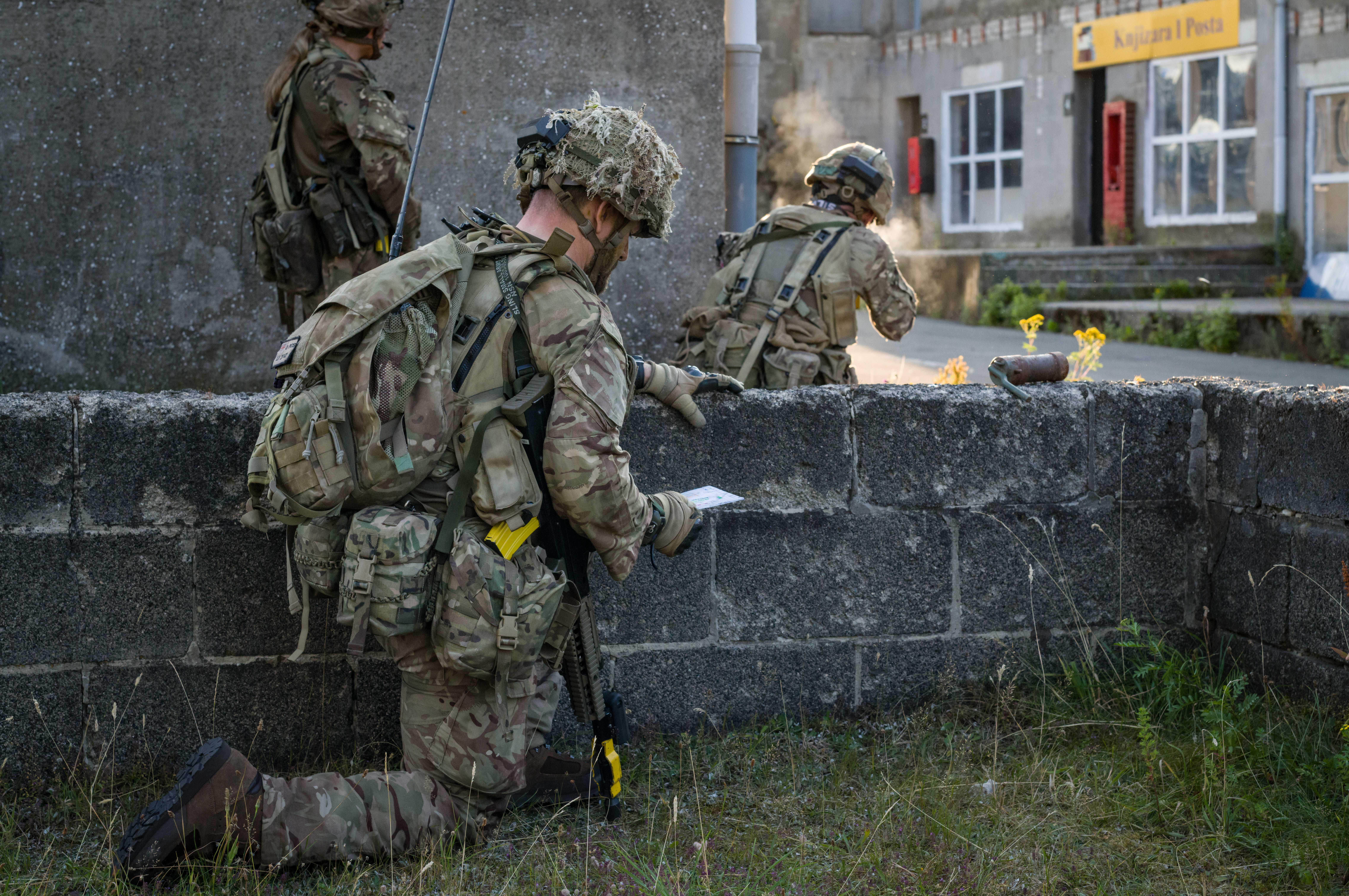 Armed personnel in camouflage uniforms and helmets, equipped with backpacks and tactical gear, are positioned behind a low stone wall. One is kneeling while holding a map, while two others are crouched and observing the area. A building with the sign 