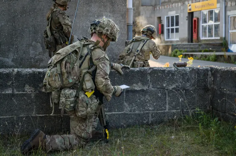 Armed personnel in camouflage uniforms and helmets, equipped with backpacks and tactical gear, are positioned behind a low stone wall. One is kneeling while holding a map, while two others are crouched and observing the area. A building with the sign