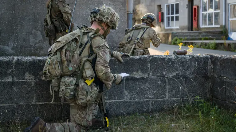 Armed personnel in camouflage uniforms and helmets, equipped with backpacks and tactical gear, are positioned behind a low stone wall. One is kneeling while holding a map, while two others are crouched and observing the area. A building with the sign