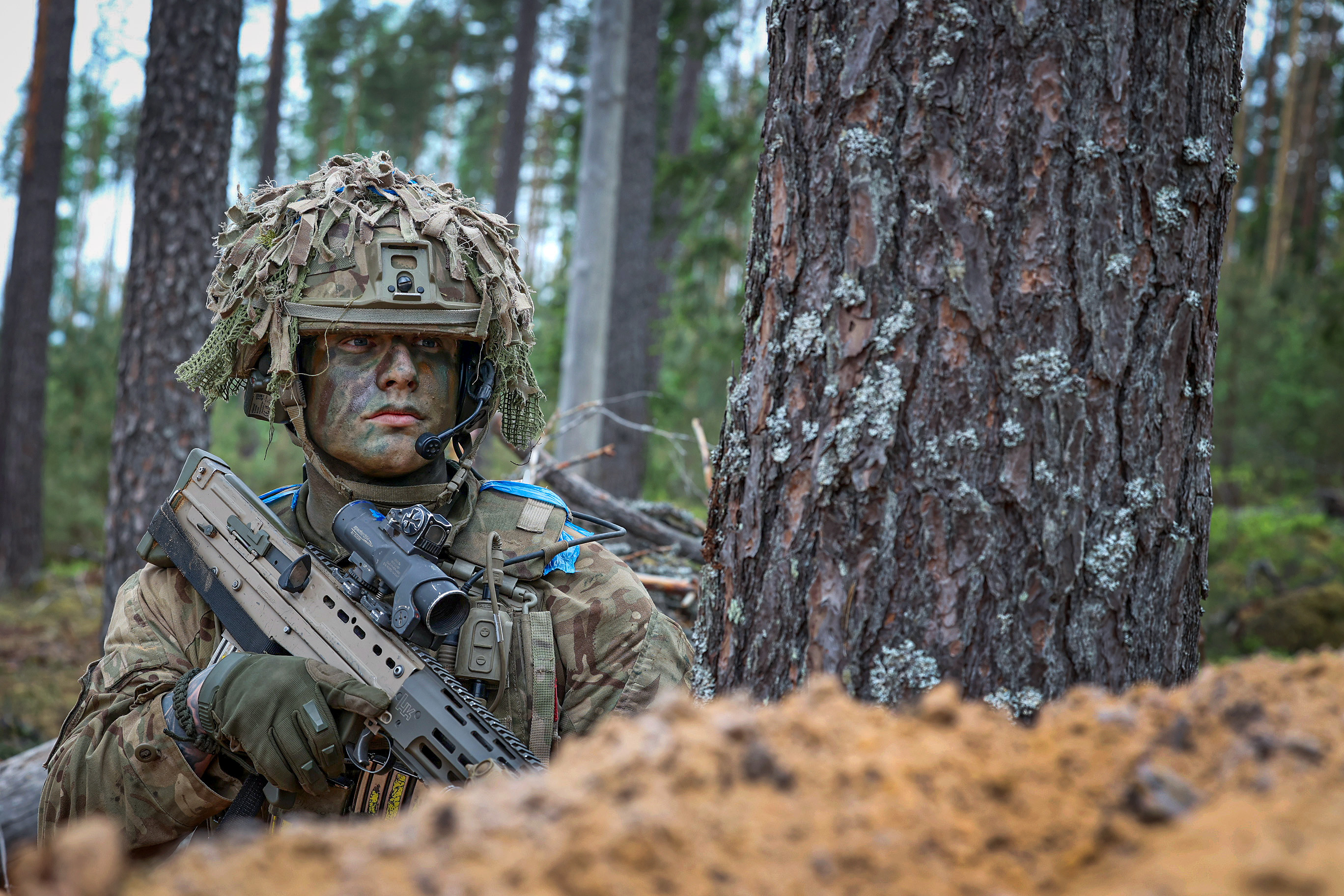A person wearing camouflage military gear and a helmet with foliage is crouched beside a tree, holding a rifle. 