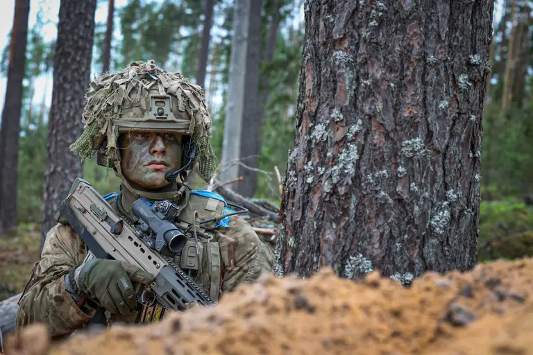 A person wearing camouflage military gear and a helmet with foliage is crouched beside a tree, holding a rifle.