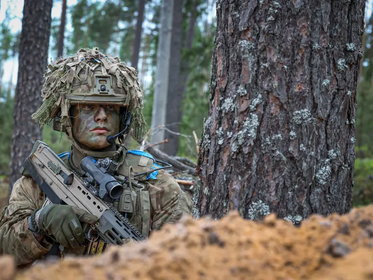 A person wearing camouflage military gear and a helmet with foliage is crouched beside a tree, holding a rifle.