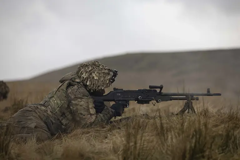 Soldier in camouflage uniform wearing body armour and helmet lays on his stomach on the firing range holding this rifle up to his eye aiming for a target. 