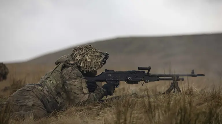 Soldier in camouflage uniform wearing body armour and helmet lays on his stomach on the firing range holding this rifle up to his eye aiming for a target.