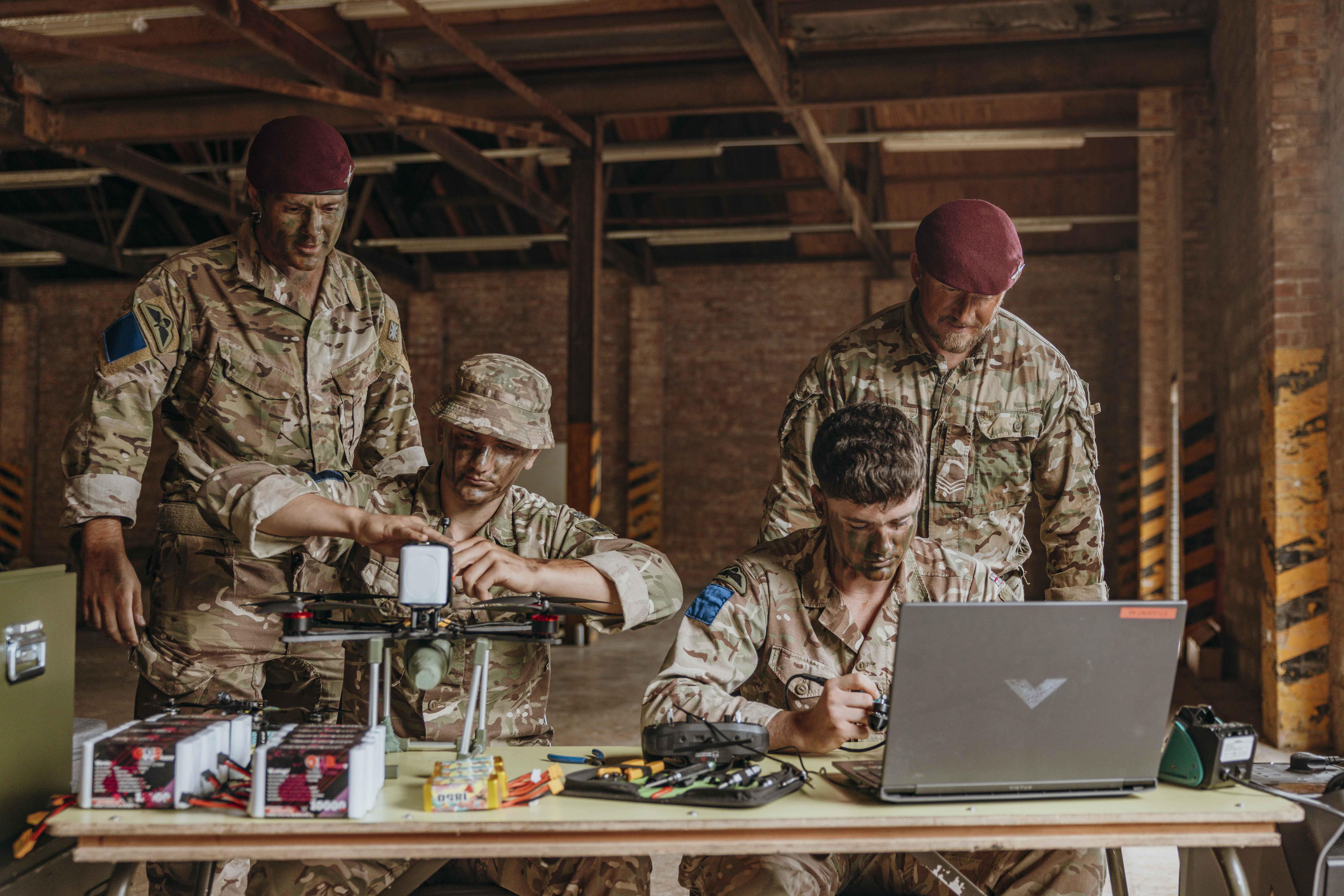 Four soldiers in camouflage uniforms work together on drone assembly inside of a warehouse with exposed brick walls.