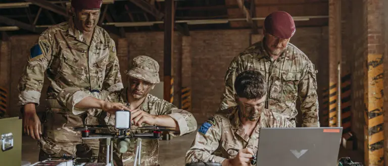 Four soldiers in camouflage uniforms work together on drone assembly inside of a warehouse with exposed brick walls.