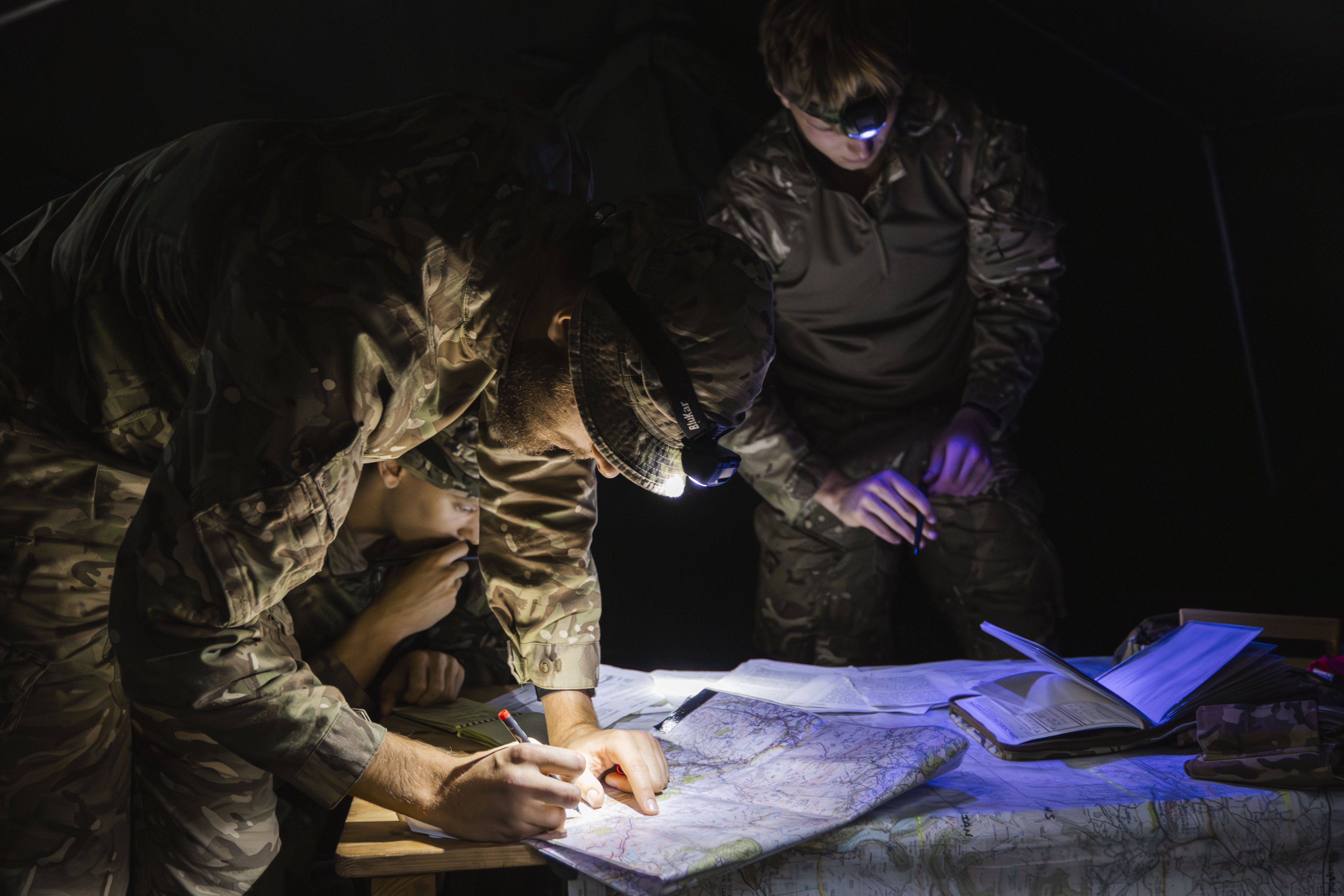 Three soldiers in camouflage gear study a map under dim light. Two bend over the map with focused expressions, while one gazes at documents.