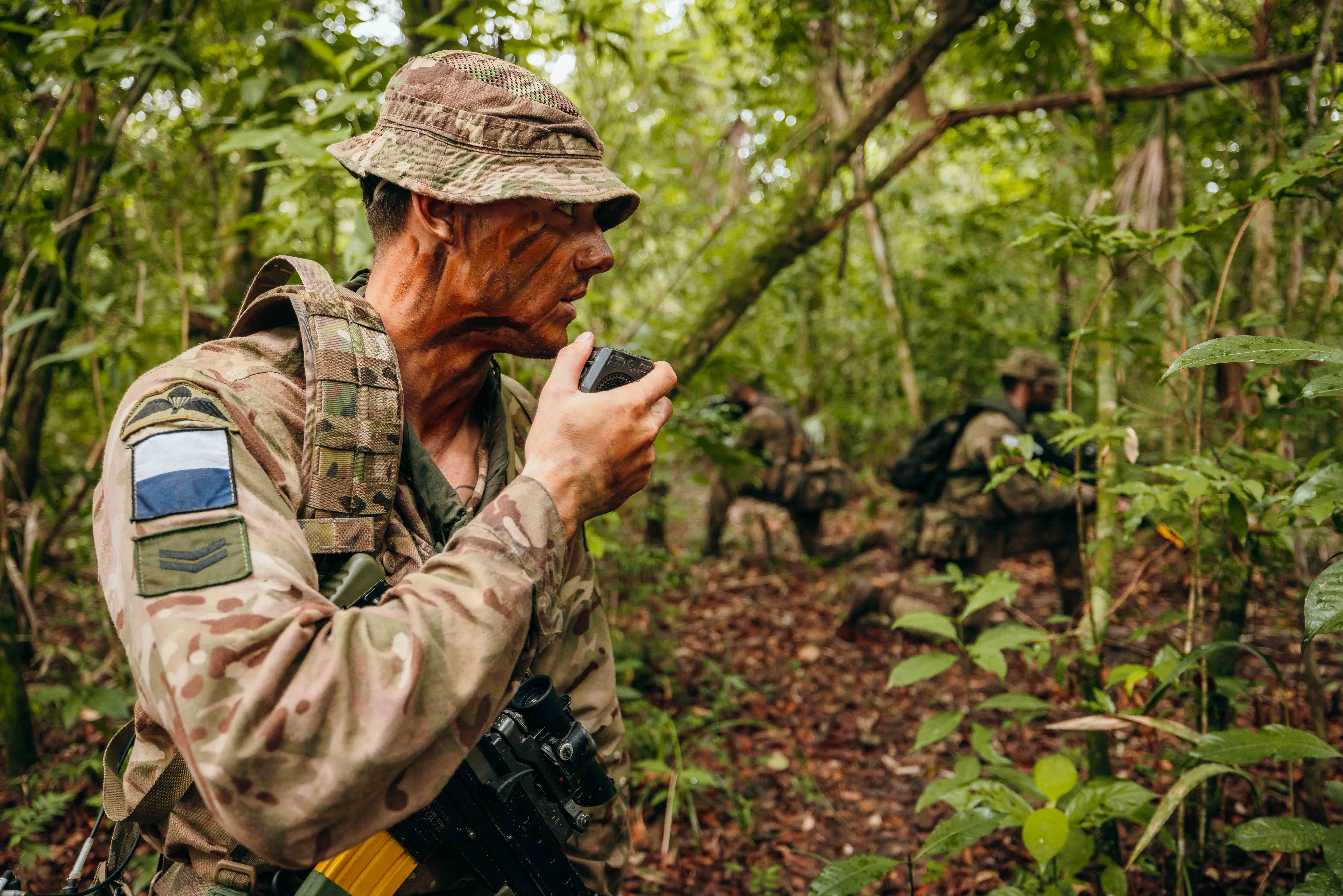 In a jungle environment, three British Army soldiers wearing camouflage uniforms, are each kneeling on one knee. The soldier on the left is talking into a radio communication device. The other soldiers are further back and holding rifles.