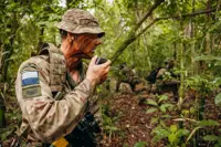 In a jungle environment, three British Army soldiers wearing camouflage uniforms, are each kneeling on one knee. The soldier on the left is talking into a radio communication device. The other soldiers are further back and holding rifles.
