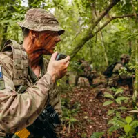 In a jungle environment, three British Army soldiers wearing camouflage uniforms, are each kneeling on one knee. The soldier on the left is talking into a radio communication device. The other soldiers are further back and holding rifles.