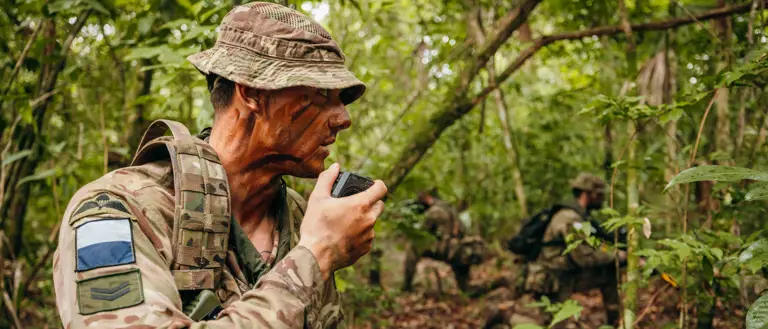 In a jungle environment, three British Army soldiers wearing camouflage uniforms, are each kneeling on one knee. The soldier on the left is talking into a radio communication device. The other soldiers are further back and holding rifles.