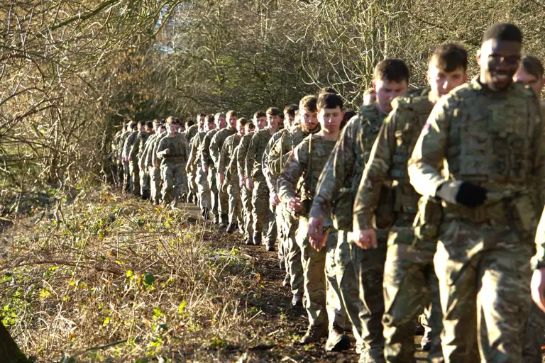 A long line of soldiers in uniform walk through a wooded area.