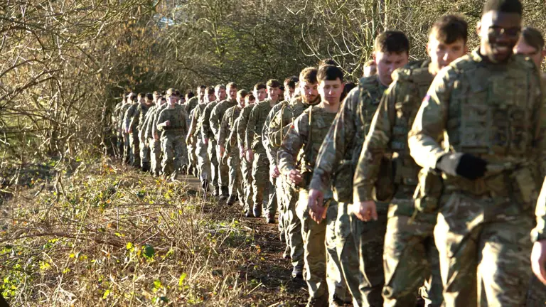 A long line of soldiers in uniform walk through a wooded area.