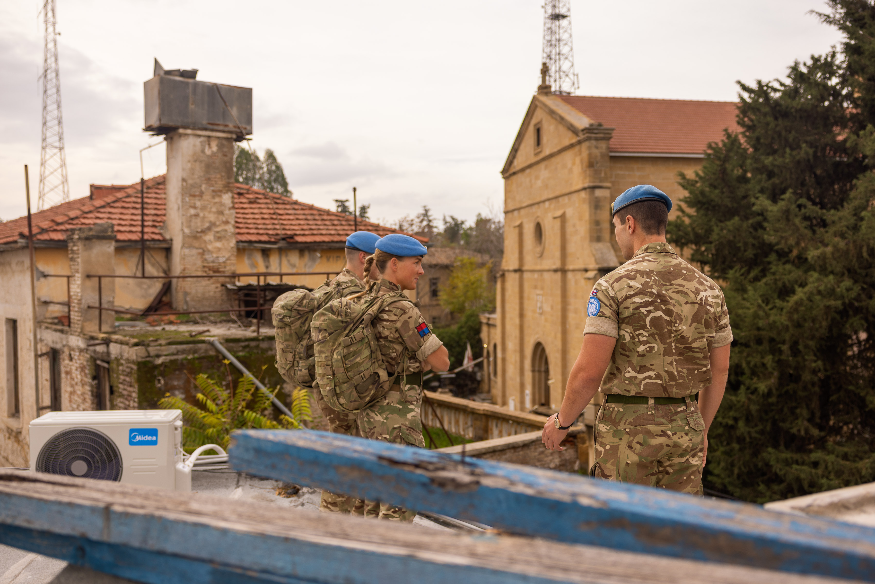Three soldiers wearing uniform and blue berets are pictured on a rooftop overlooking a church. 