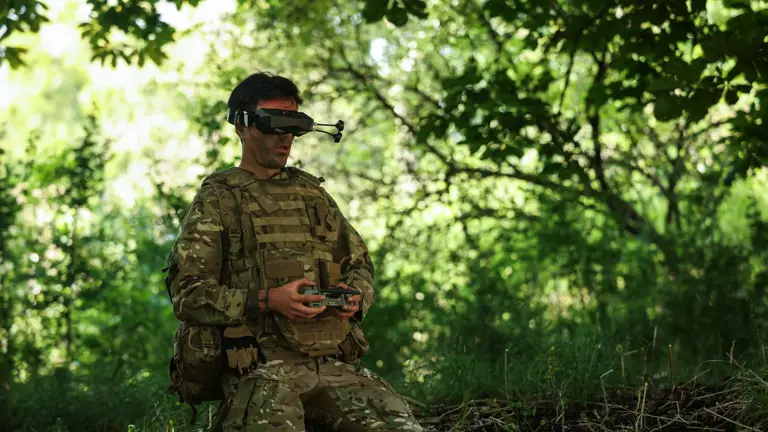 A man in uniform kneels whilst flying a drone in a wooded area.