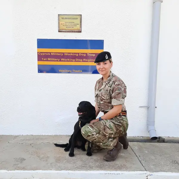 A person in military uniform kneels beside a black dog in front of a sign for the Cyprus Military Working Dog Troop. The scene is calm and professional.