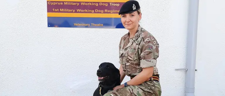 A person in military uniform kneels beside a black dog in front of a sign for the Cyprus Military Working Dog Troop. The scene is calm and professional.