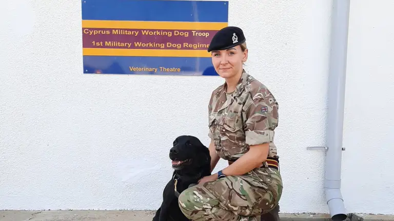 A person in military uniform kneels beside a black dog in front of a sign for the Cyprus Military Working Dog Troop. The scene is calm and professional.