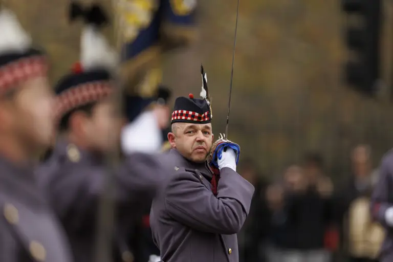 Uniformed soldier in ceremonial attire raising a sword during a formal event outdoors.