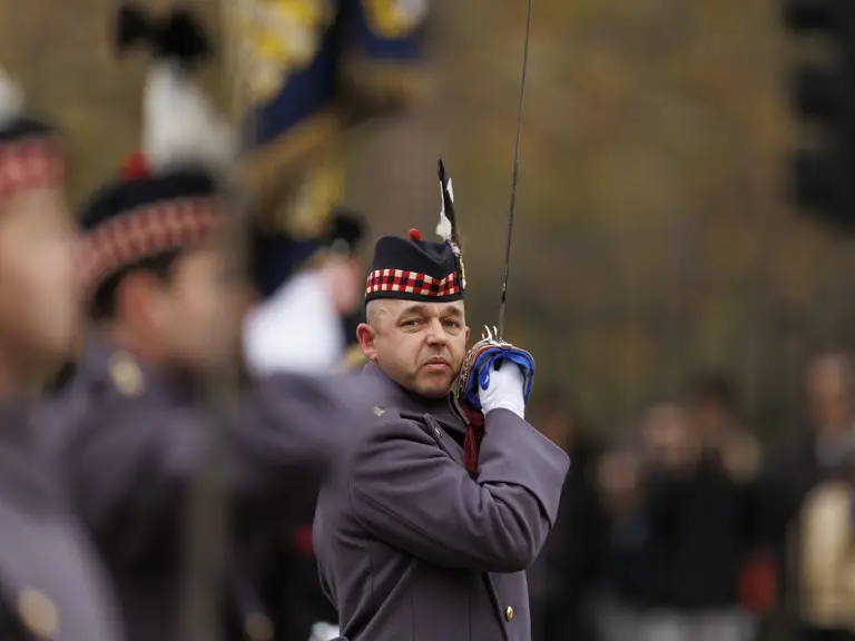 Uniformed soldier in ceremonial attire raising a sword during a formal event outdoors.