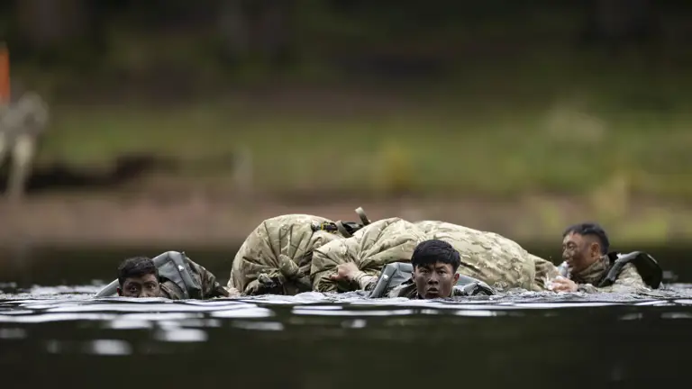 Individuals in military uniforms are wading through water, carrying a large pack. They appear focused and determined. The setting is outdoors, with a forested background.