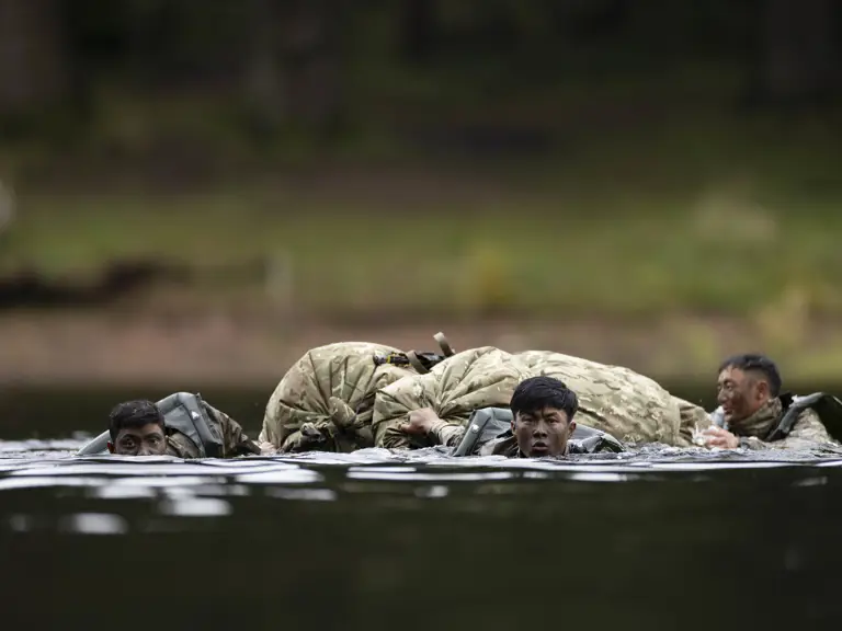 Individuals in military uniforms are wading through water, carrying a large pack. They appear focused and determined. The setting is outdoors, with a forested background.