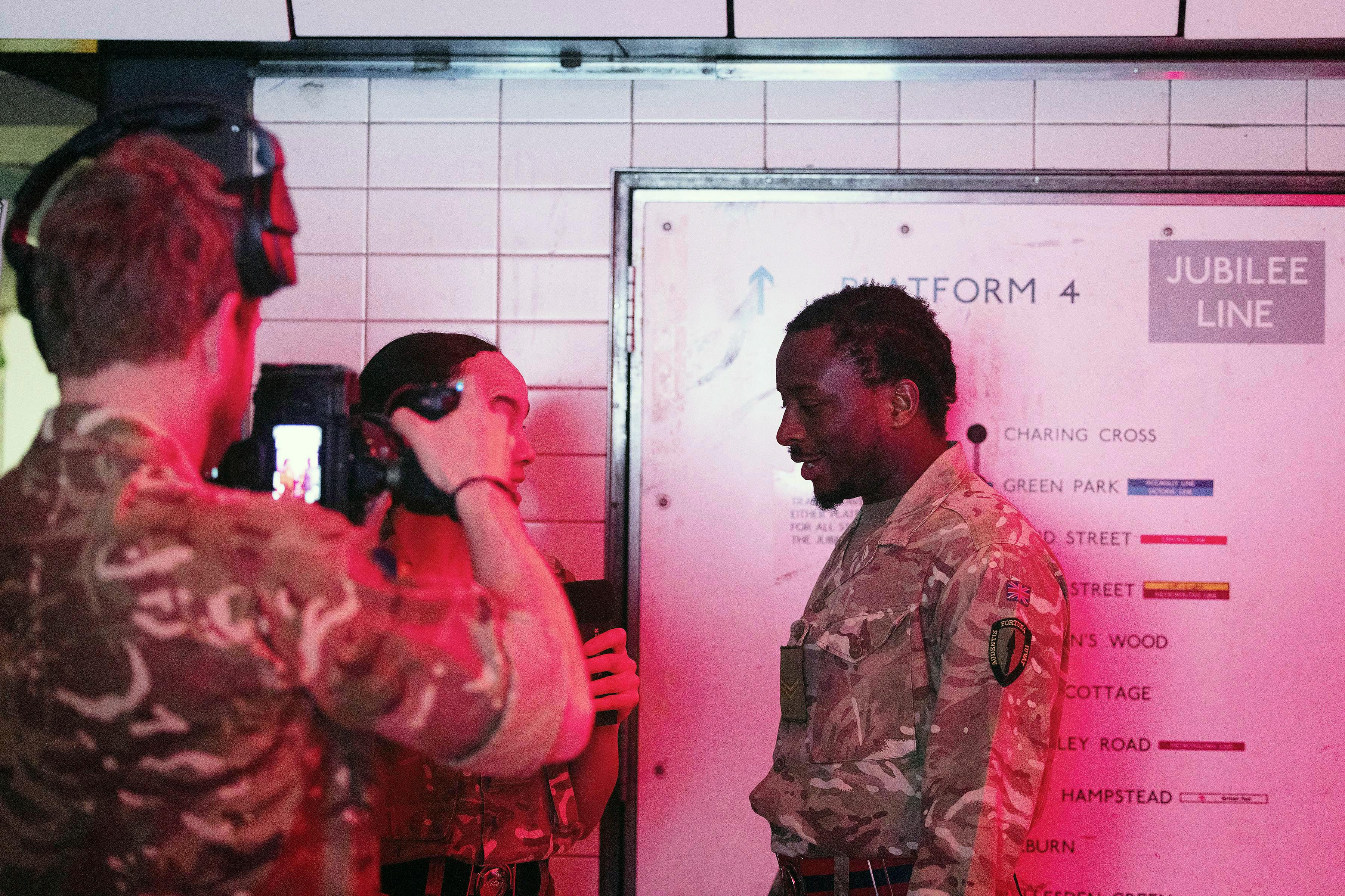 A group of individuals in camouflage uniforms stand on a subway platform beneath a 